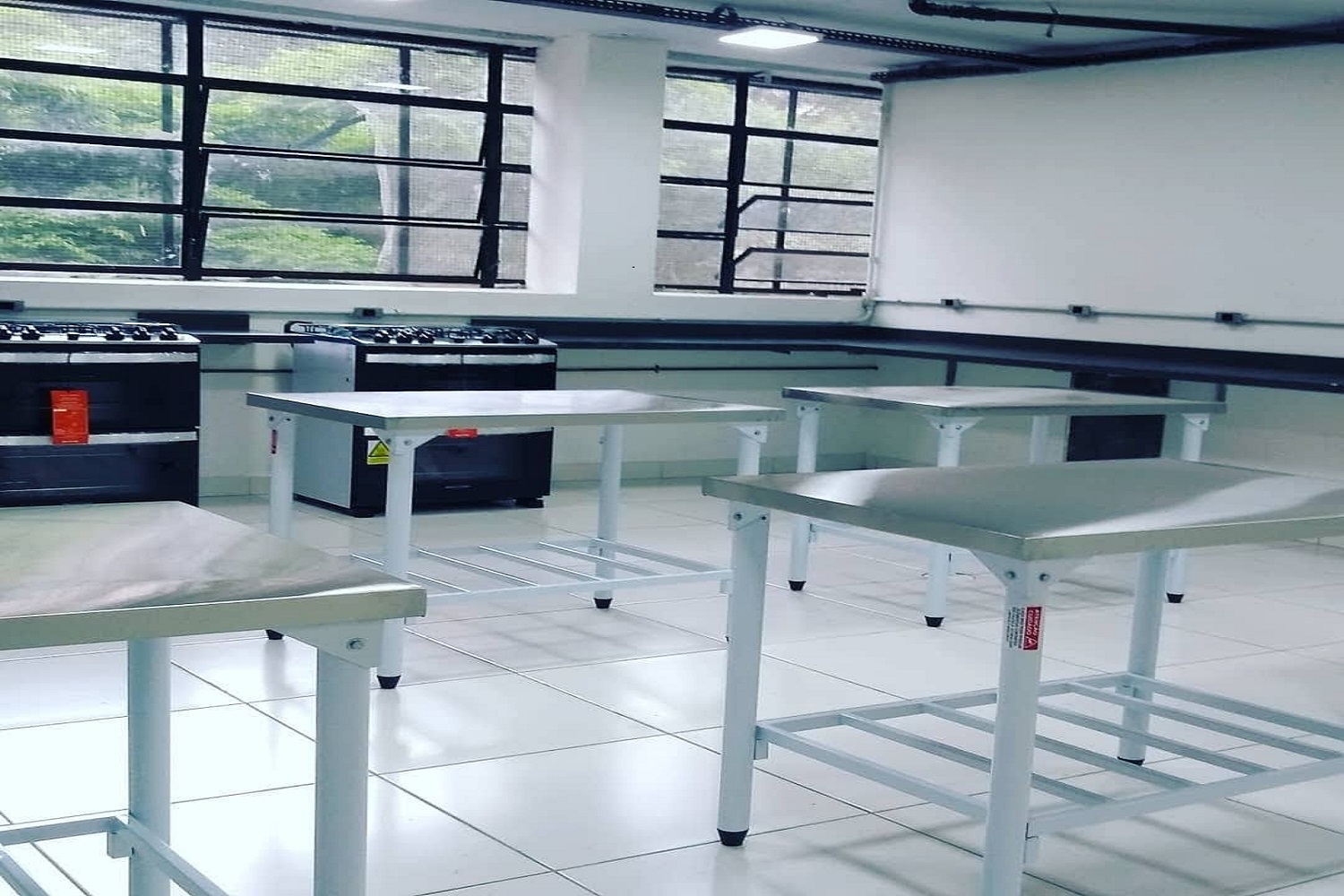 Cozinha do Cisarte com quatro bancadas móveis de inox, balcão de granito, dois fogões novos com etiquetas e janelas grandes ao fundo. 